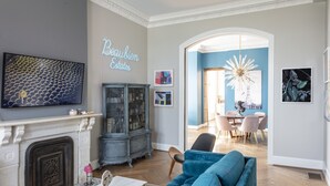 A living room with a fireplace, a blue sofa, a television, and a dining area in the background. - NEW! Brush Park Brilliance! Walk->Stadiums DT Luxe 3BR (Detroit)