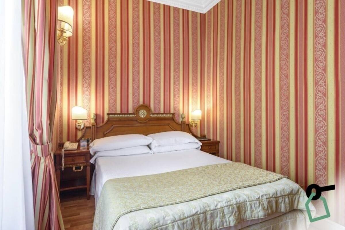 Une chambre d’hôtel avec un grand lit, deux tables de chevet, une petite table et une lampe fixée au mur.