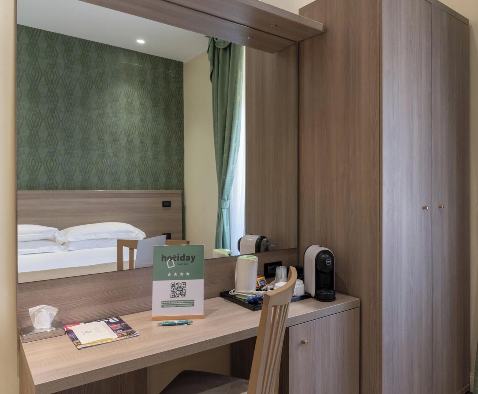 A hotel room with a bed, a desk with a sign, a coffee machine, and a cabinet.