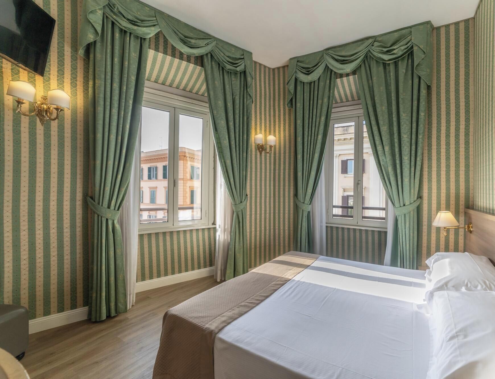 A hotel room with a bed, two windows with curtains, a wall-mounted TV, and a wall sconce.
