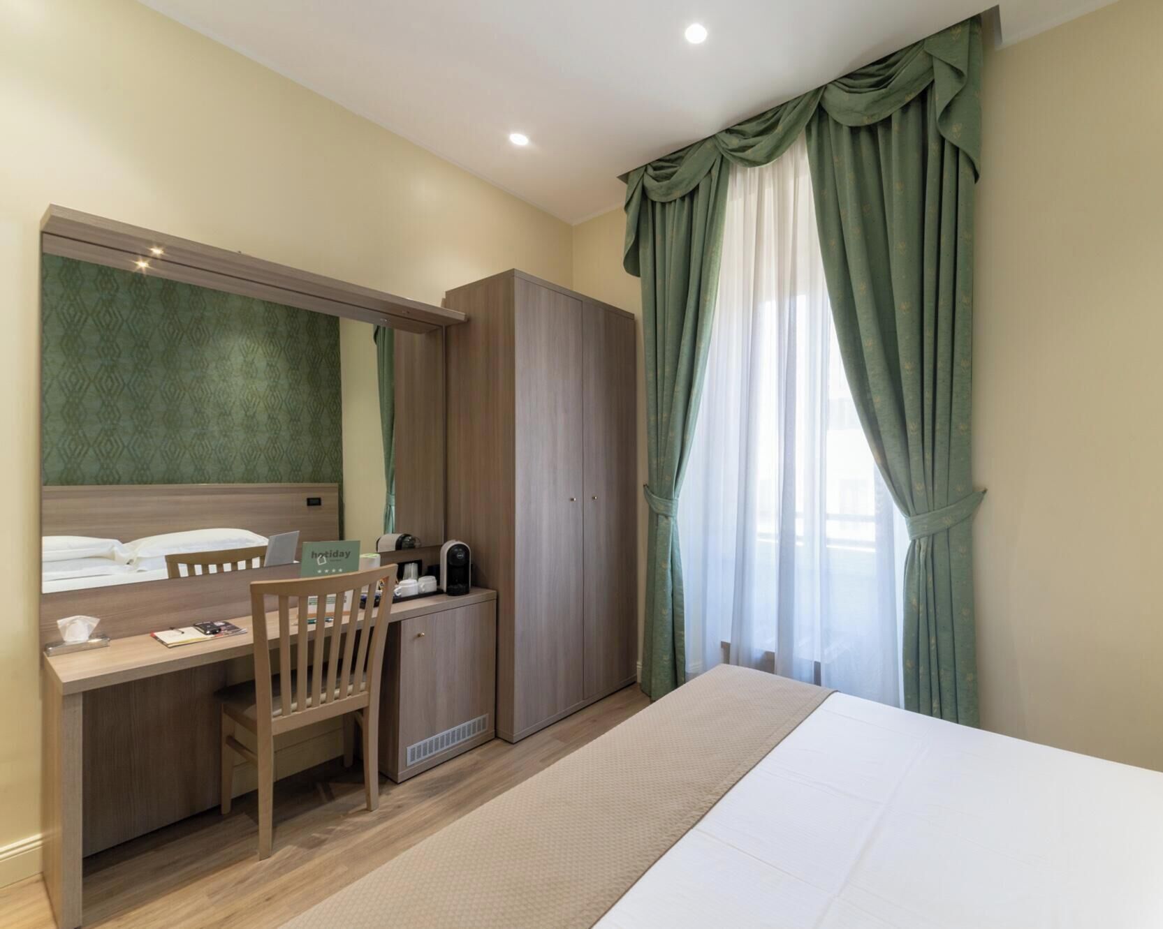 Una habitación de hotel con una cama, un escritorio con silla, un armario y una ventana con cortinas.