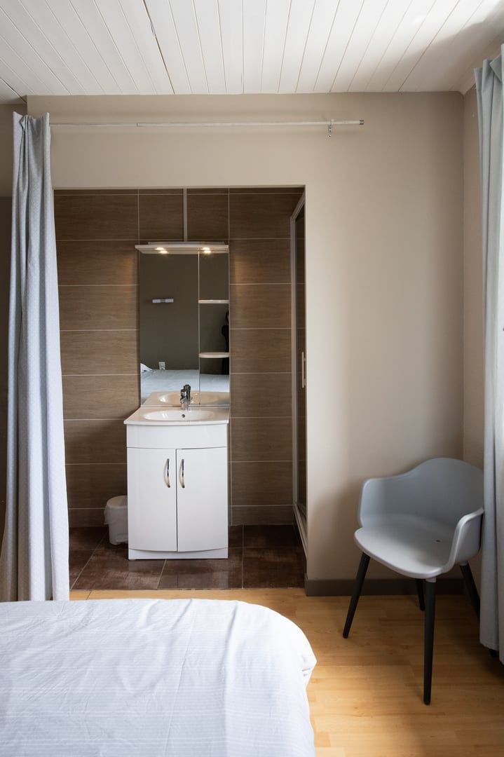 A modern bedroom with a bed, a white cabinet with a mirror, a grey chair, and a wooden floor.