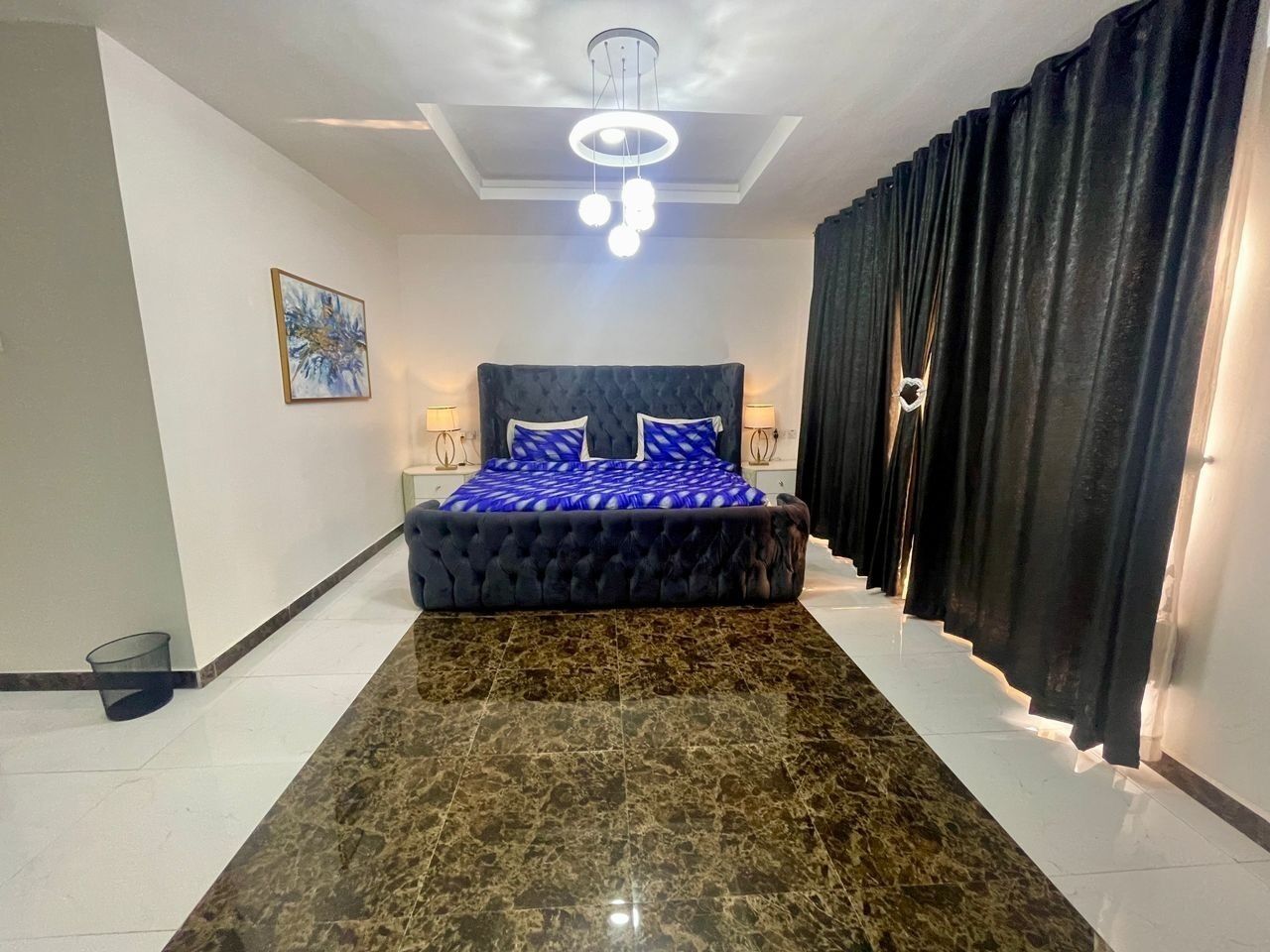 A bedroom with a tufted headboard, a large bed with blue bedding, a marble floor, and a ceiling light fixture.