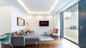 Un salon moderne avec un canapé gris, des coussins colorés, une table basse en verre et un téléviseur à écran plat affichant le logo « NETFLIX ».