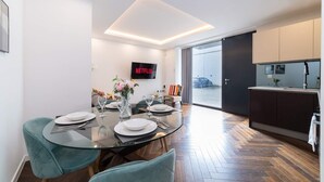 Un espace de salle à manger moderne avec une table en verre, des chaises et un vase de fleurs. Un téléviseur à écran plat est fixé au mur, et on aperçoit une cuisine au fond.