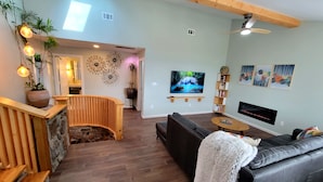A modern living room with a stone fireplace, wooden staircase, and a ceiling fan.