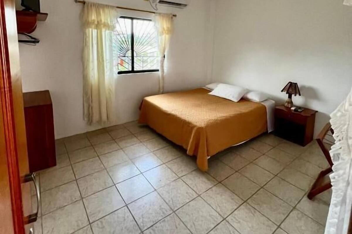 Une petite chambre propre, équipée d’un lit, d’une télévision, d’une fenêtre avec des rideaux et d’un appareil de climatisation.