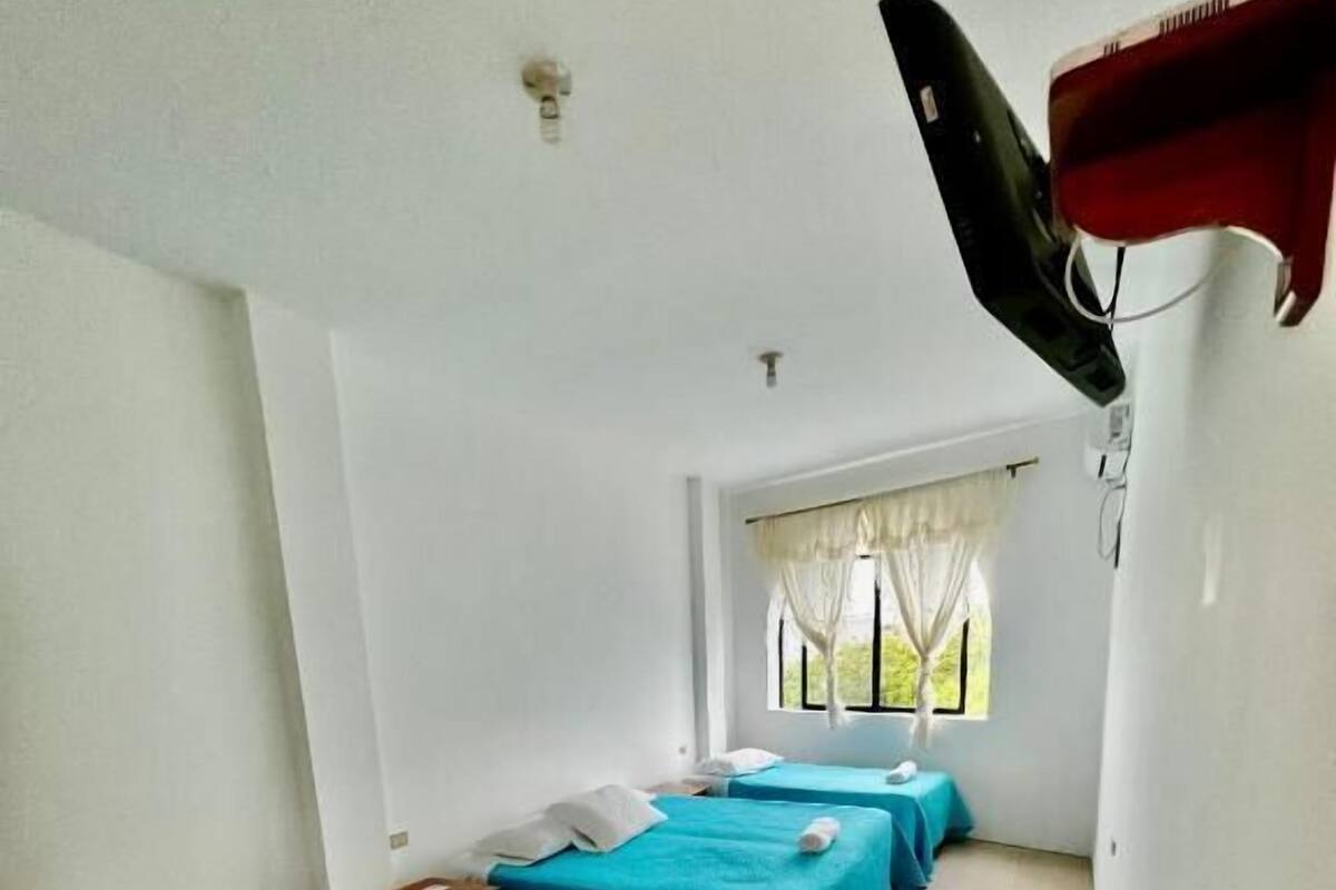Une chambre d’hôtel avec trois lits simples, chacun recouvert d’une couverture bleue et agrémenté d’oreillers blancs. Une fenêtre offre une vue sur la verdure environnante.