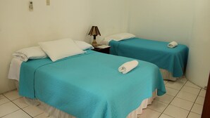 Habitación de hotel con dos camas individuales, cada una con una colcha color turquesa y almohadas blancas. Hay una mesita de noche con una lámpara entre las camas.