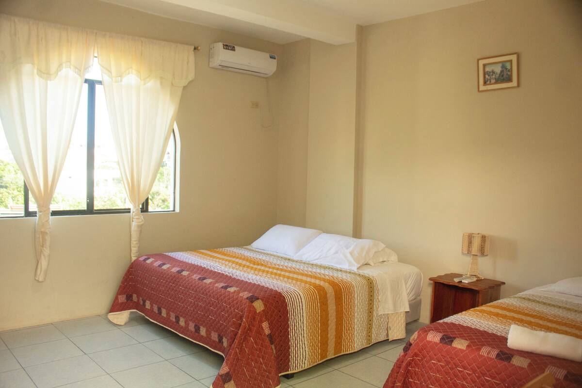 Une chambre avec deux lits, une fenêtre avec des rideaux, un appareil de climatisation et une petite table avec une lampe.