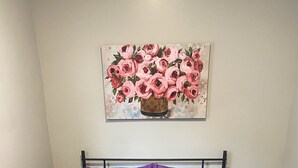 Una cama con colcha de patchwork morada y almohadas a juego, un cabecero de metal negro, un cuadro de flores rosadas en la pared y una mesita de noche con una lámpara blanca.