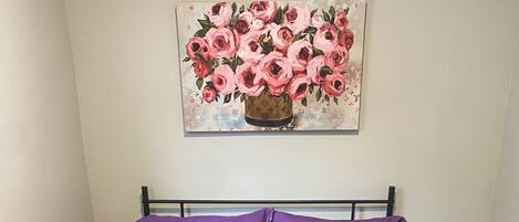 A bed with a purple quilted bedspread and matching pillows, a black metal headboard, a wall-mounted painting of pink flowers, and a bedside table with a white lamp.
