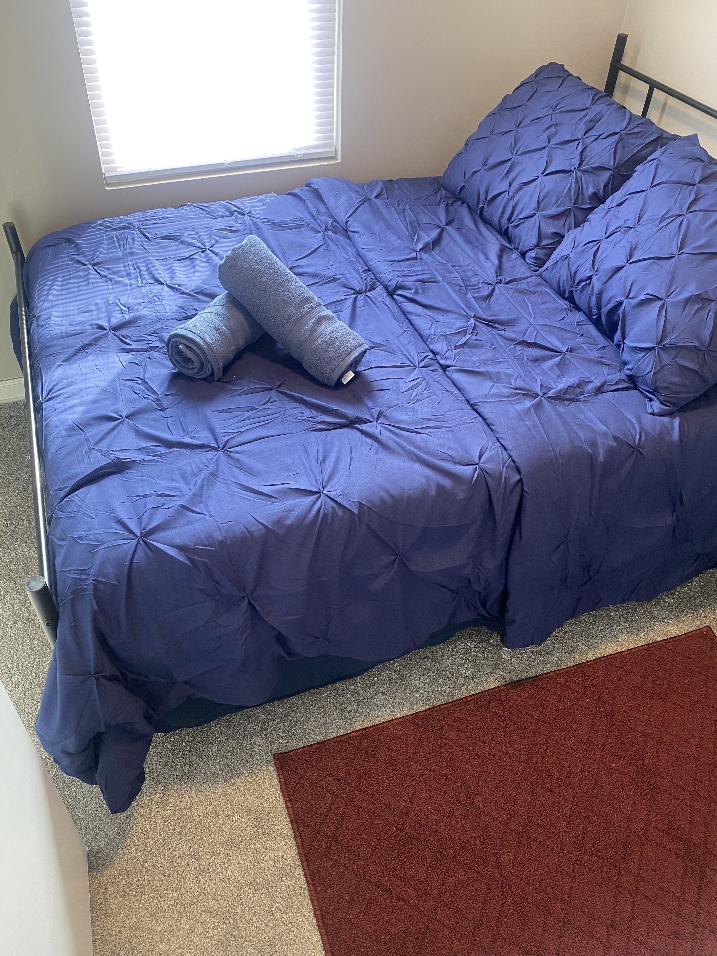 Un lit avec une couette bleue et des serviettes enroulées, un tapis marron et une fenêtre avec des stores.