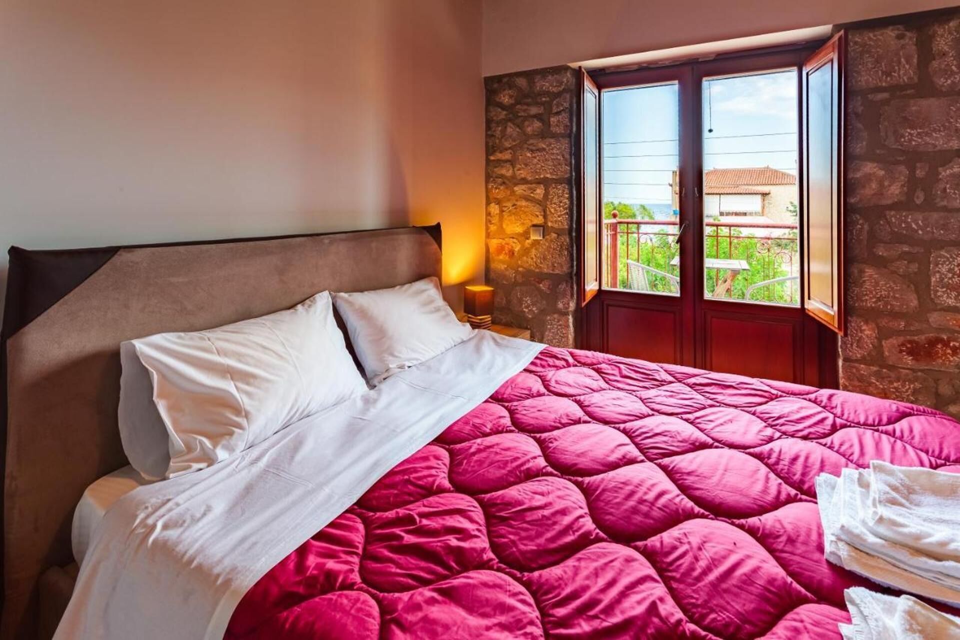 A bed with a pink quilt and white pillows, a stone wall, and a window with a view of greenery.