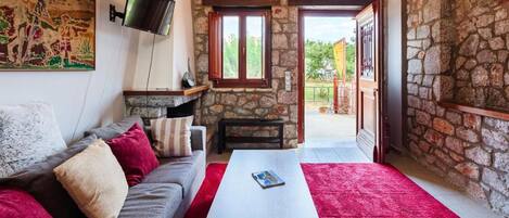A cozy living room with a stone wall, a flat-screen TV mounted on the wall, a grey sofa with red and white cushions, a white coffee table, and a red carpet.