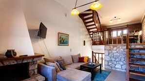 A living room with a stone fireplace, a grey sofa, a wooden staircase, and a TV mounted on the wall.