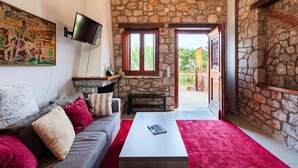 A cozy living room with a stone wall, a flat-screen TV mounted on the wall, a grey sofa with red and white cushions, a white coffee table, and a red carpet.