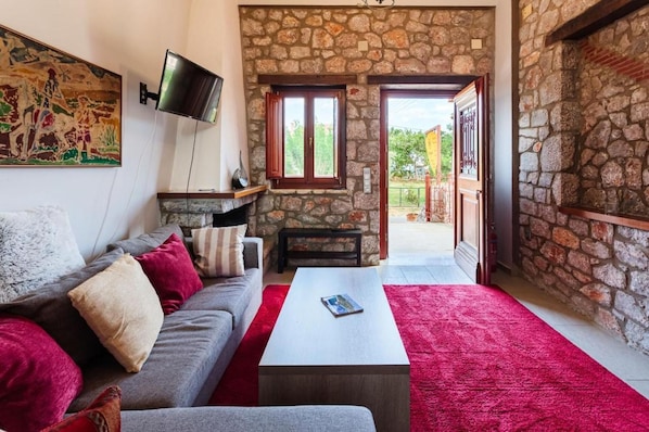 A cozy living room with a stone wall, a flat-screen TV mounted on the wall, a grey sofa with red and white cushions, a white coffee table, and a red carpet.
