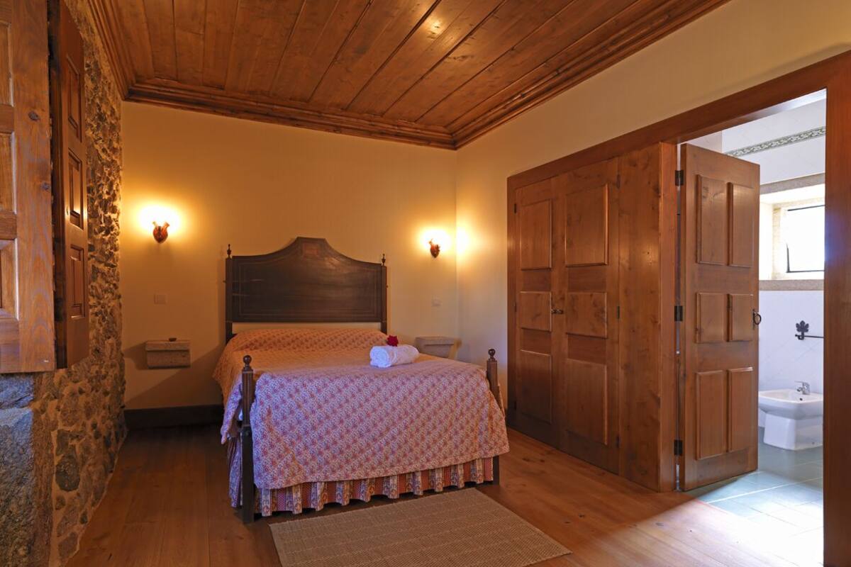 A bedroom with a wooden ceiling, a bed with a patterned bedspread, two wall-mounted lights, a wooden wardrobe, and a bathroom visible through an open door.