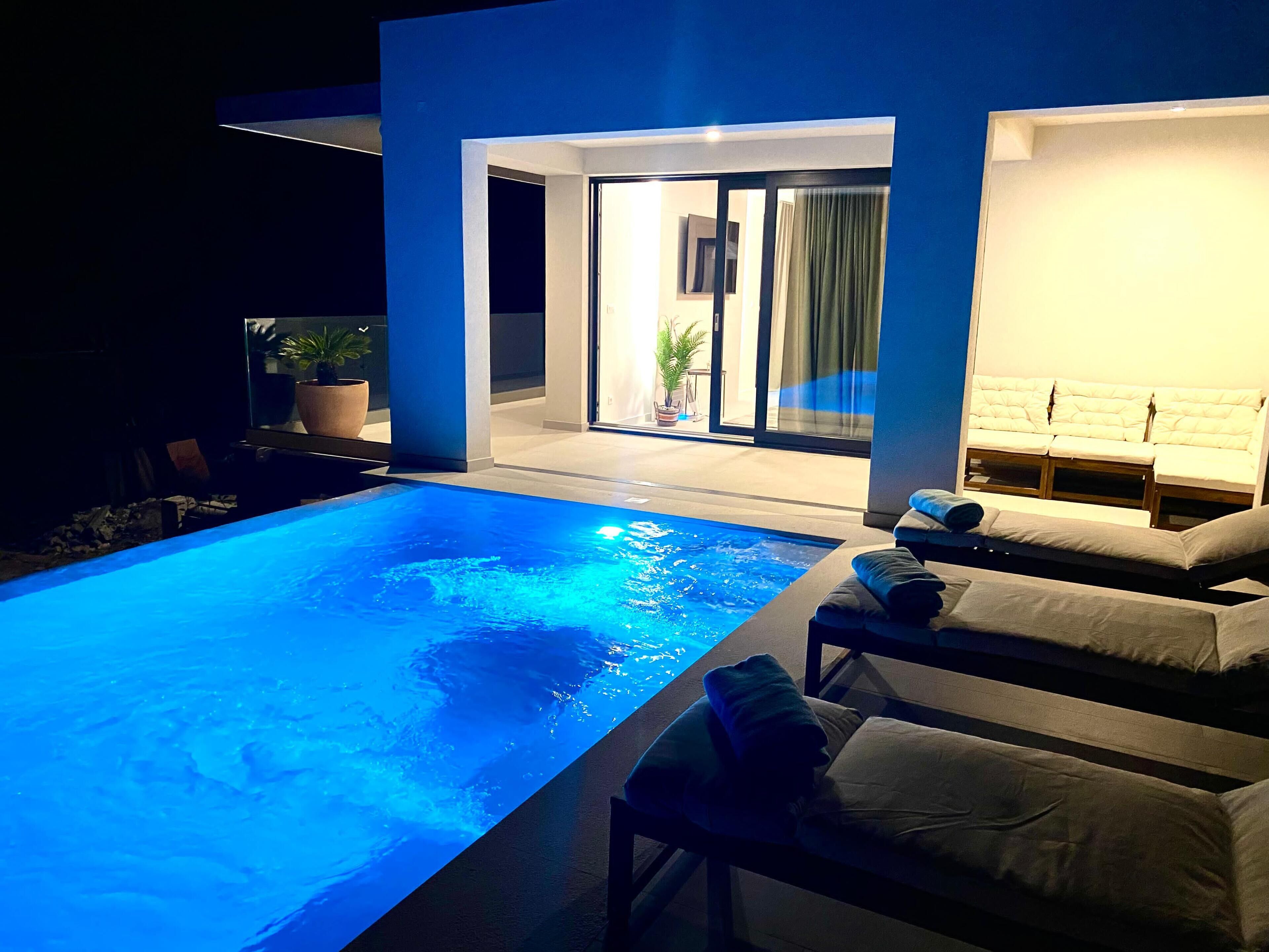 A modern indoor pool with a clear blue water feature, adjacent to a seating area with cushioned loungers and a glass door leading to a bright room.