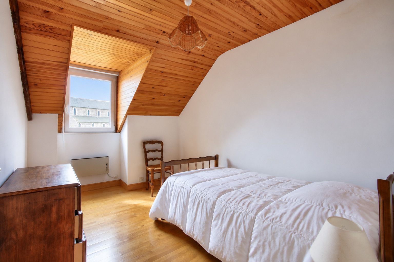 Ein Schlafzimmer mit schrägem Dach, einem Bett, einem Sessel, einer Kommode und einem Fenster mit Blick auf Gebäude.