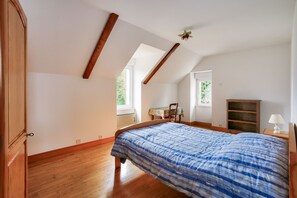 Ein Schlafzimmer mit schrägen Wänden, Holzbalken, einem Bett mit blauen und weißen Mustern, einem Nachttisch aus Holz, einem Sessel, einem kleinen Tisch mit Lampe und einem Holzschrank.
