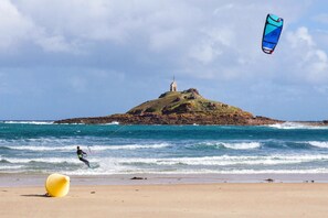 Ein Strand mit einer Person, die Windsurfen geht, einem Leuchtturm auf einem Hügel und einem gelben Ball im Sand.