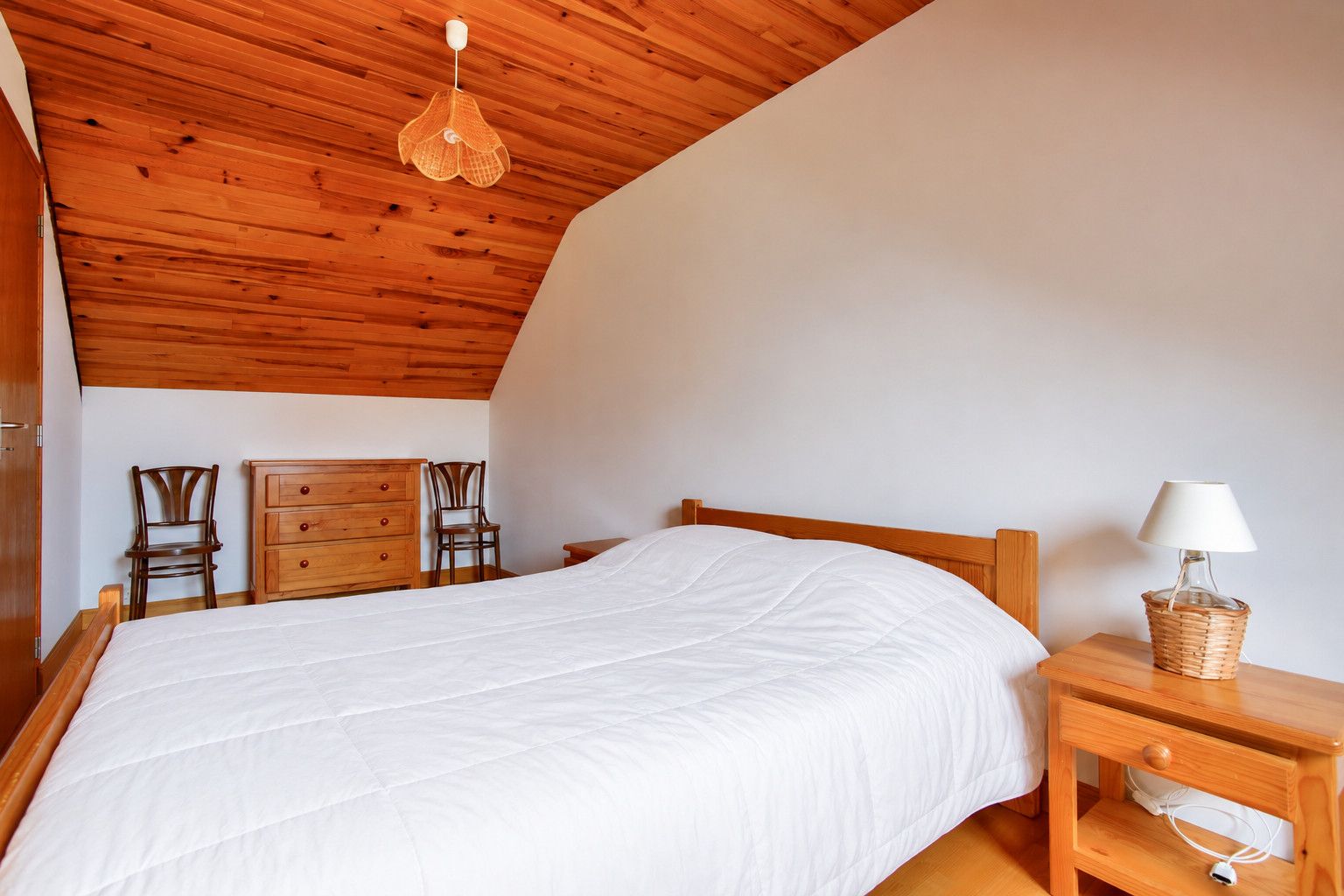 Quarto com teto de madeira, cama com roupa de cama branca, duas cadeiras de madeira, cômoda de madeira e mesa de cabeceira com abajur e cesta.