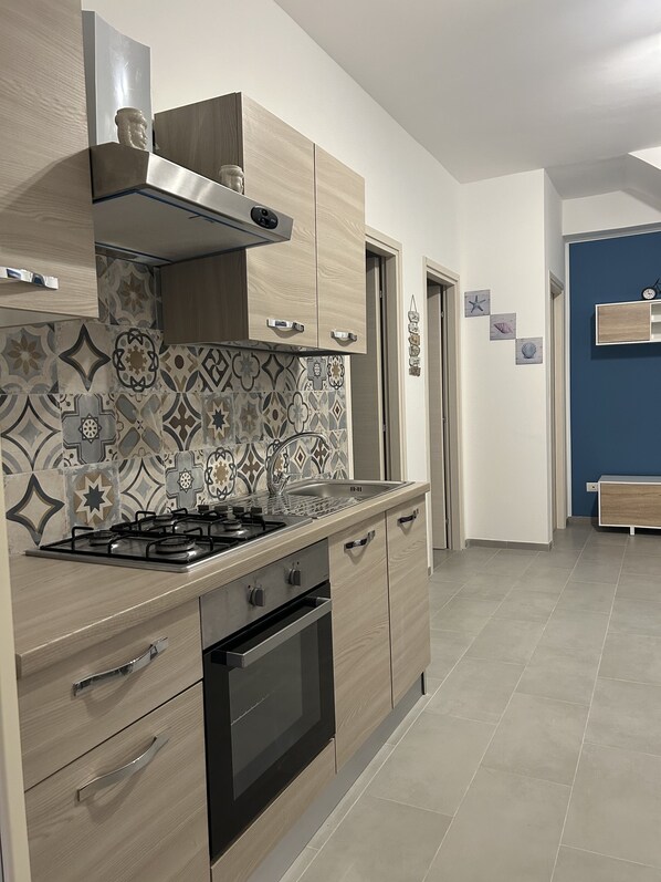 A modern kitchen with a gas stove, oven, and stainless steel sink. There are wooden cabinets and a patterned backsplash. - SUMMER HOUSE (Mazzeo)