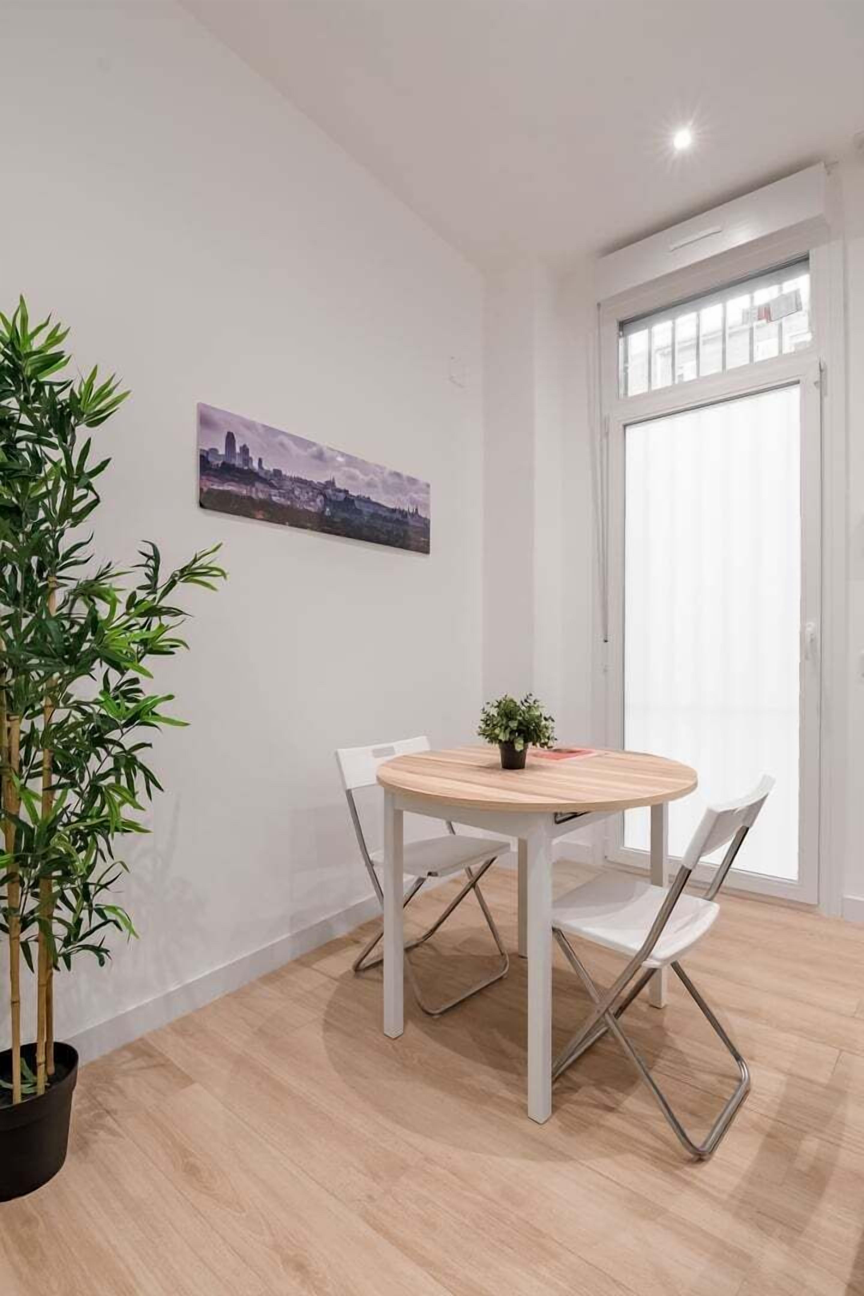 Una habitación con una mesa redonda de madera, dos sillas blancas, una planta en maceta y un cuadro de un paisaje urbano en la pared.