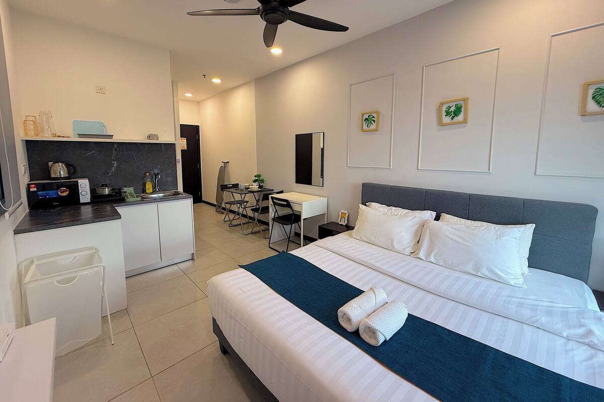 A hotel room with a bed, a desk, a kitchenette, and a ceiling fan.
