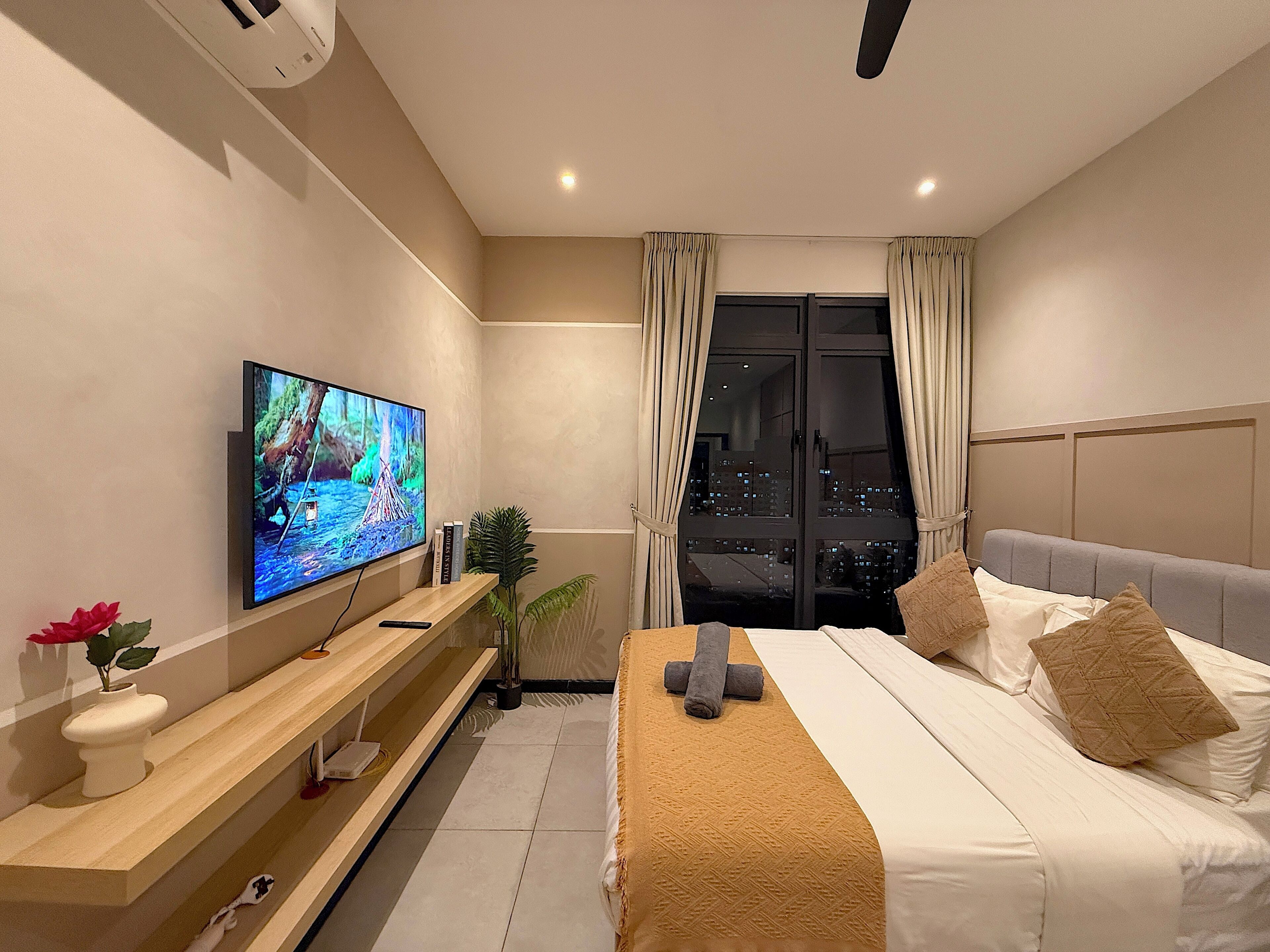 A modern hotel room with a large flat-screen TV, a bed with beige bedding, a wooden console table, and a potted plant.