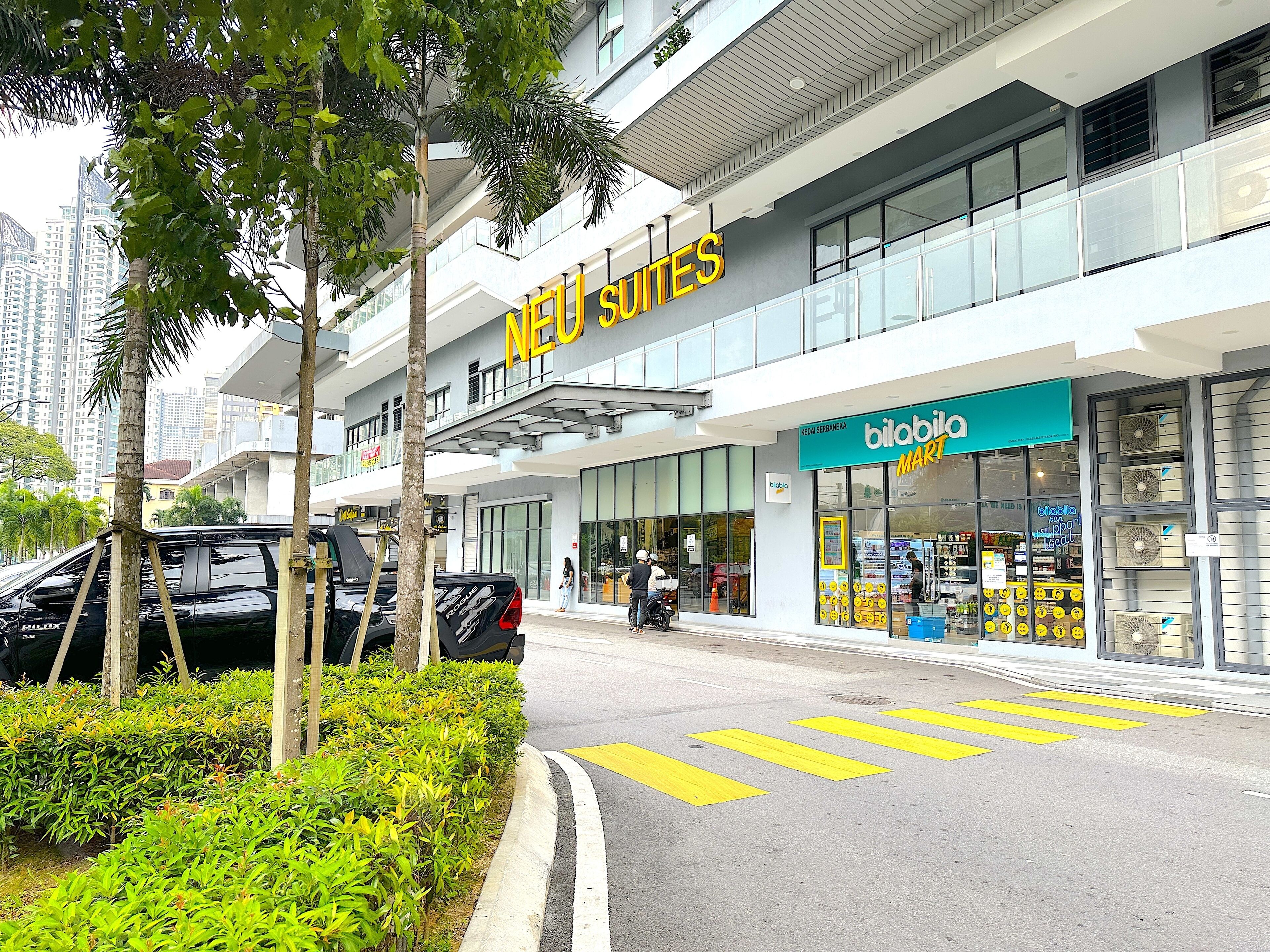 A modern hotel with a prominent 'NEU Suites' sign, a 'bilabia MART' store across the street, and a clear pedestrian crossing.