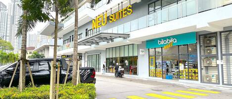 A modern hotel with a prominent 'NEU Suites' sign, a 'bilabia MART' store across the street, and a clear pedestrian crossing.