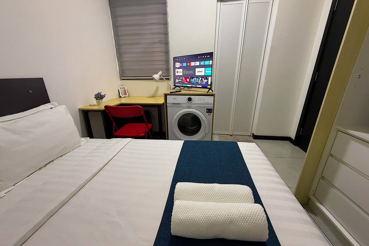 A hotel room with a bed, a desk, a washing machine, and a computer monitor.