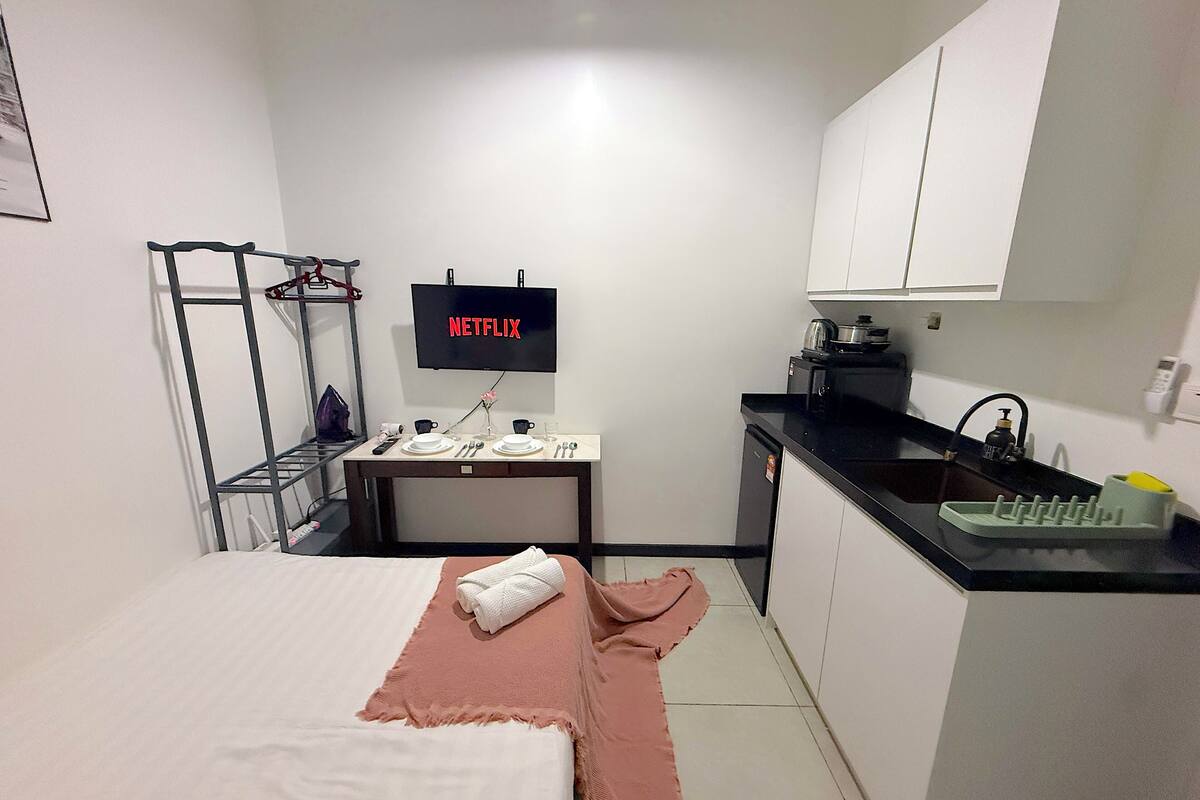 A compact hotel room with a bed, a small kitchenette, a TV displaying 'NETFLIX', and a metal shelf unit.