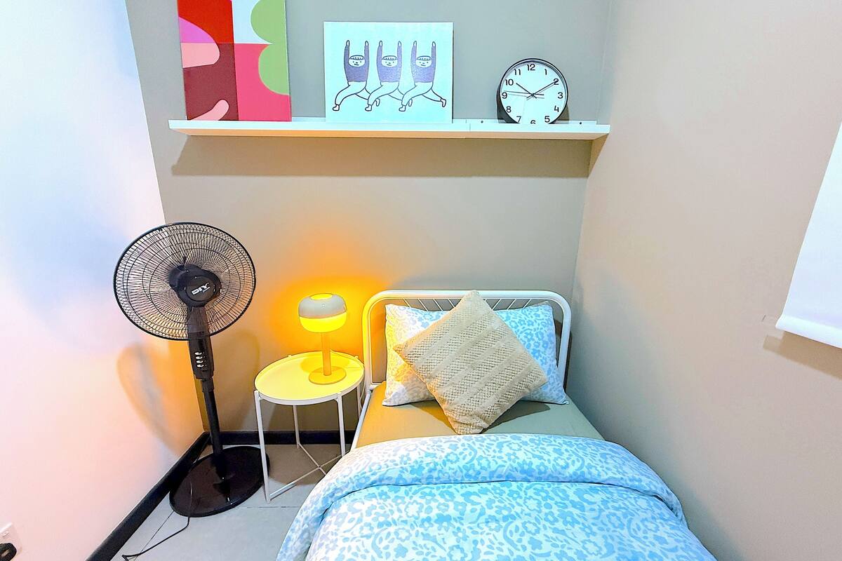 A small bedroom with a bed, a bedside table with a lamp, a wall clock, and a floor fan.