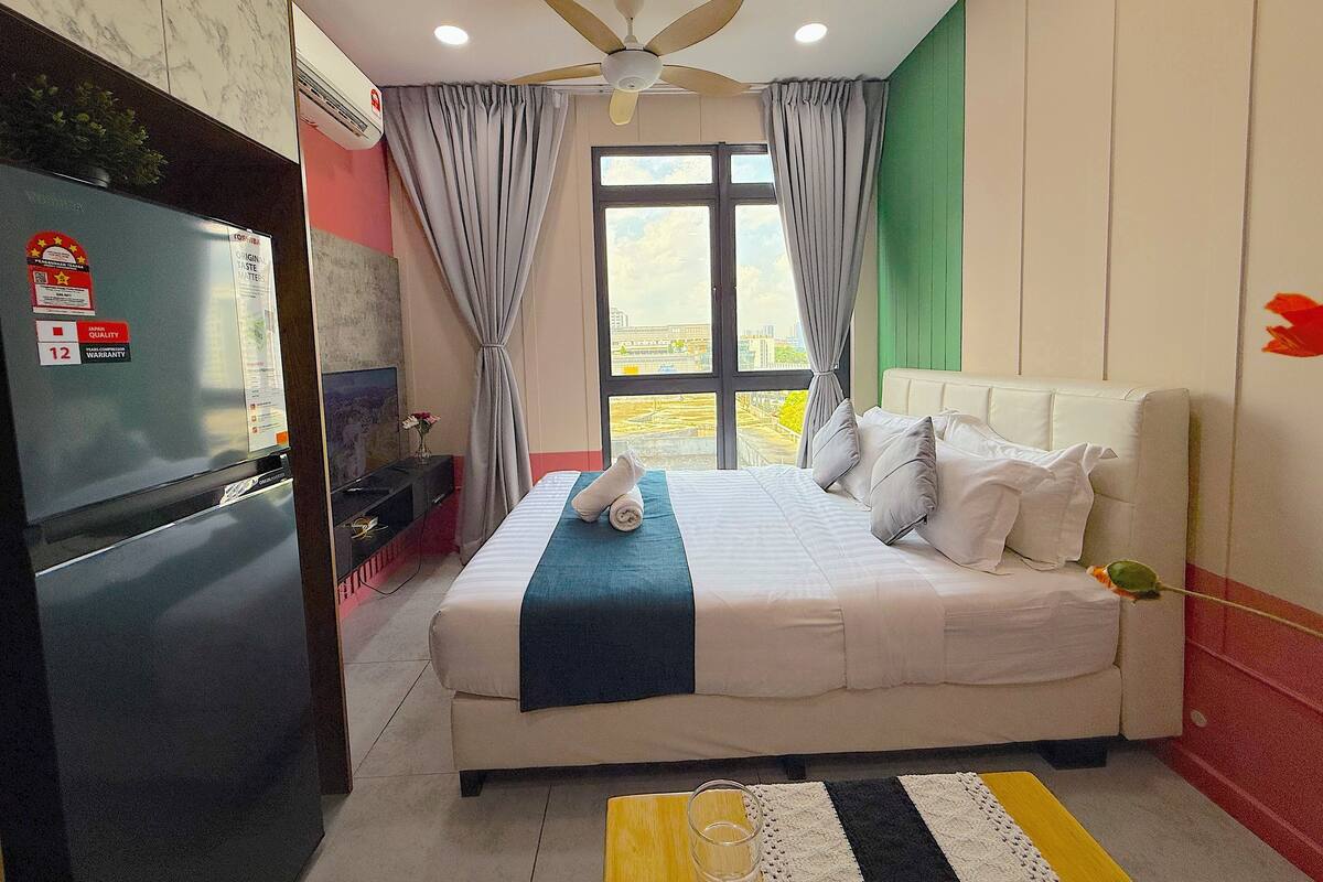 A hotel room with a bed, a ceiling fan, a window with curtains, a refrigerator, and a view of the city.