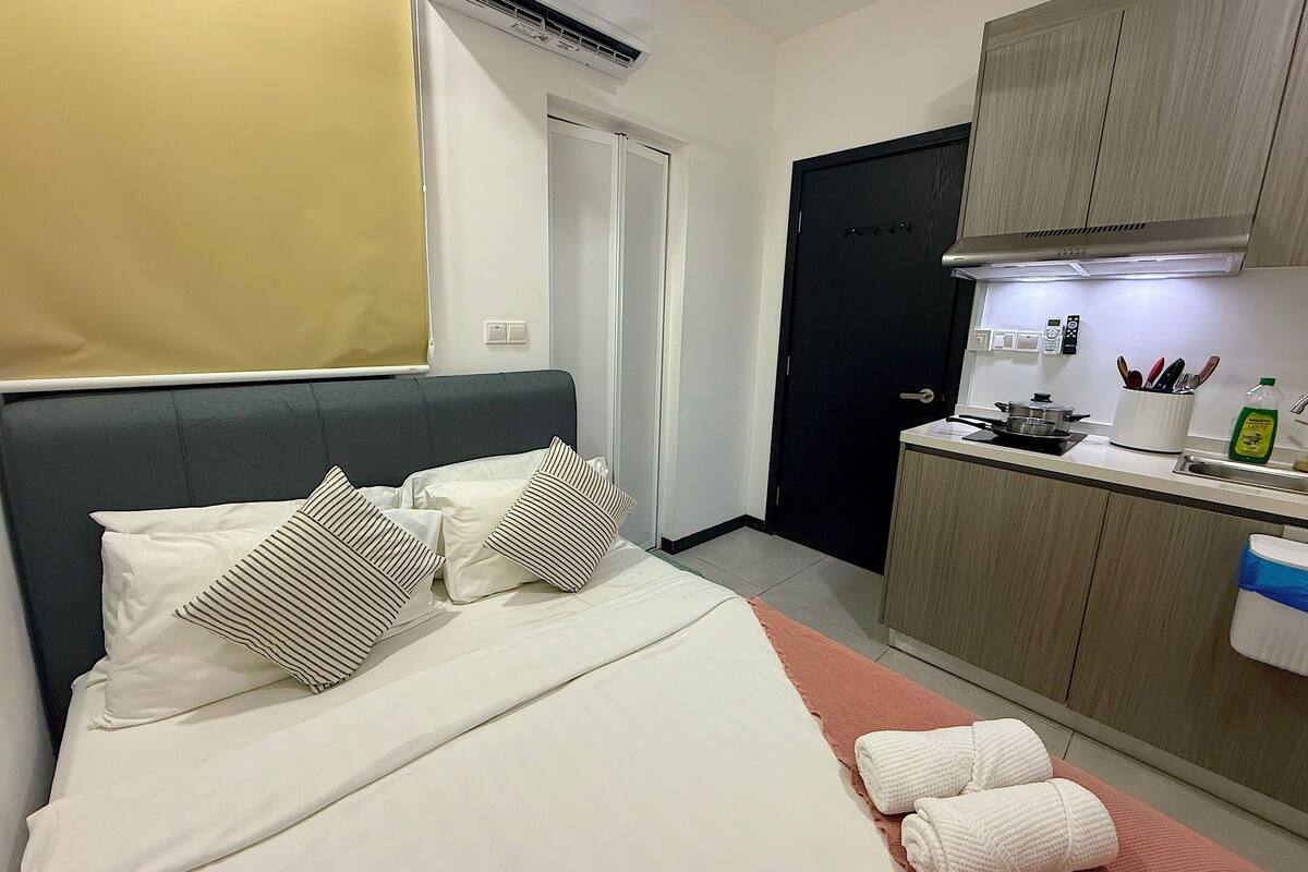 A compact hotel room with a bed, bedside table, and a kitchenette area.