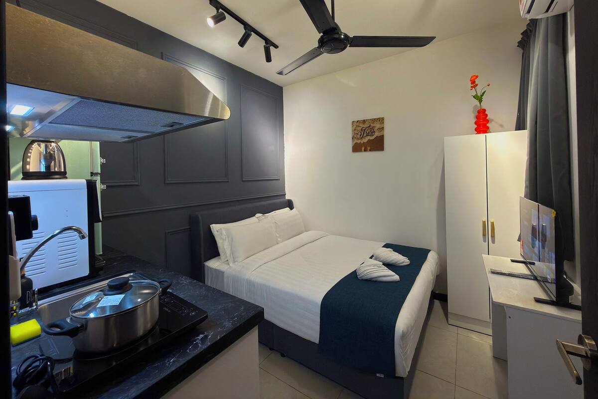 A compact hotel room with a bed, a small kitchenette, a ceiling fan, and a TV.