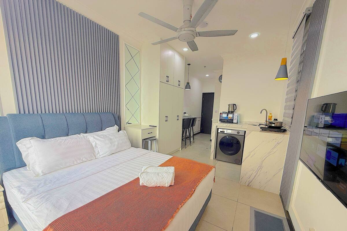 A bedroom with a bed, a desk, a washing machine, and a TV.