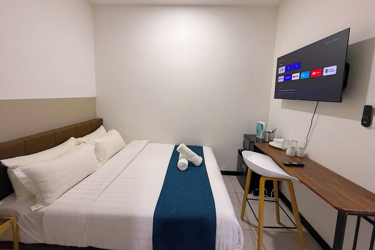 A hotel room with a bed, a desk, a TV mounted on the wall, and a remote control on the desk.