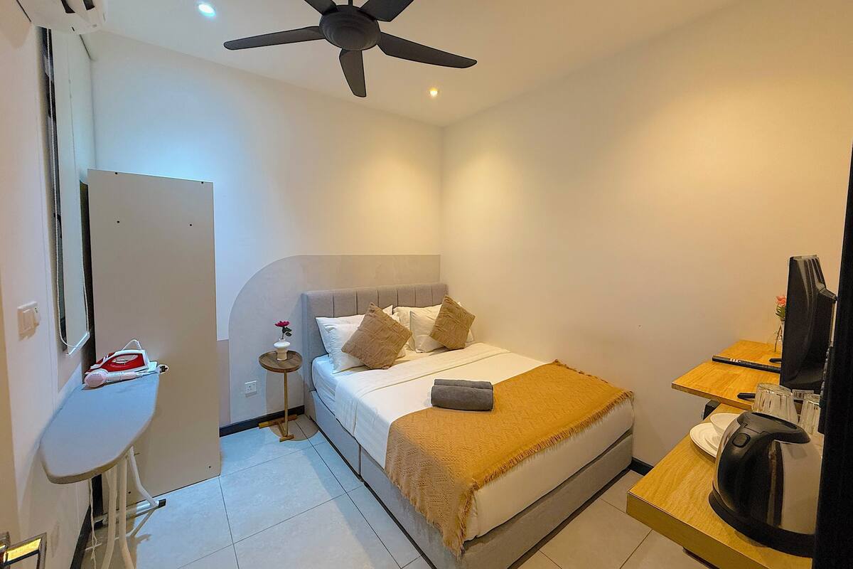 A hotel room with a bed, a desk with a computer, a ceiling fan, and a small table with a kettle.