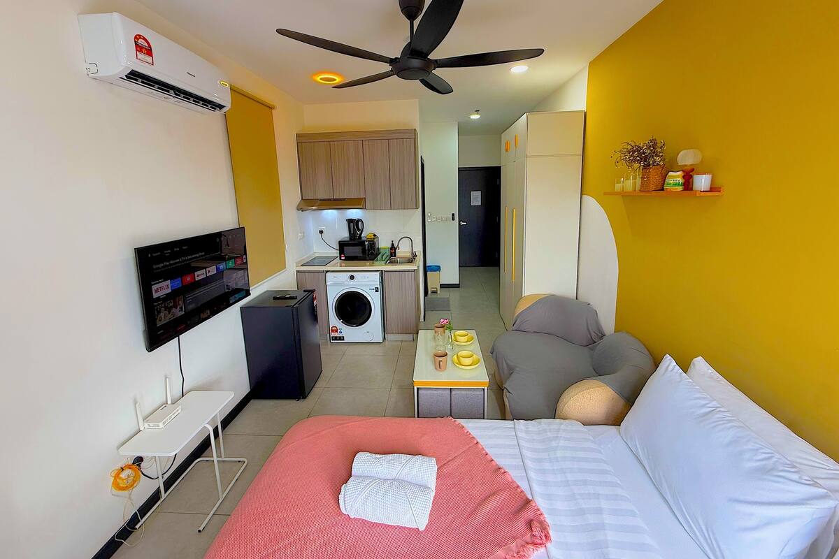 A compact hotel room with a bed, a desk, a chair, a washing machine, a kitchenette, and a ceiling fan.