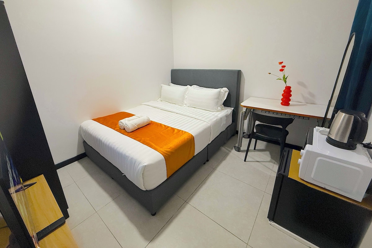 A hotel room with a bed, a desk, a chair, a small table with a red vase, and a microwave.