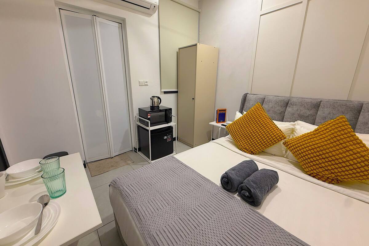 A hotel room with a bed, a bedside table, a microwave, a kettle, and a small table with a book and a cup.
