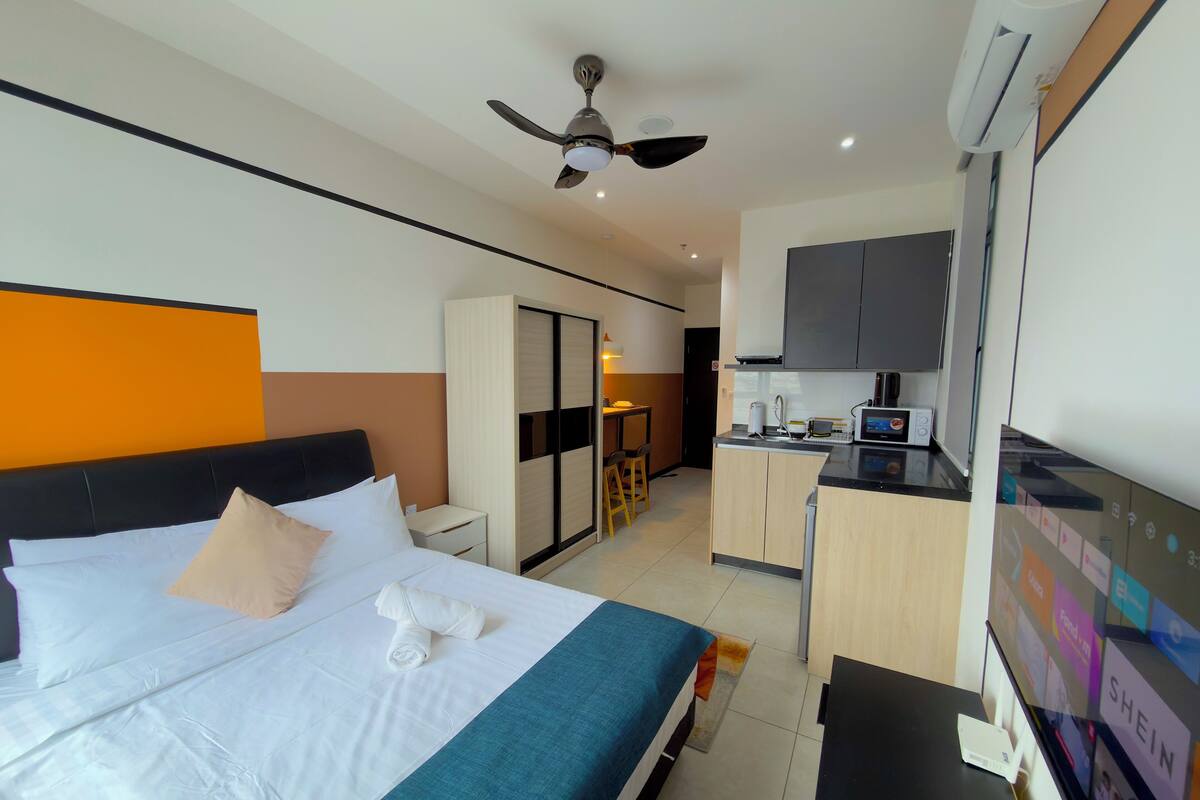 A hotel room with a bed, a desk, a kitchenette, and a ceiling fan.