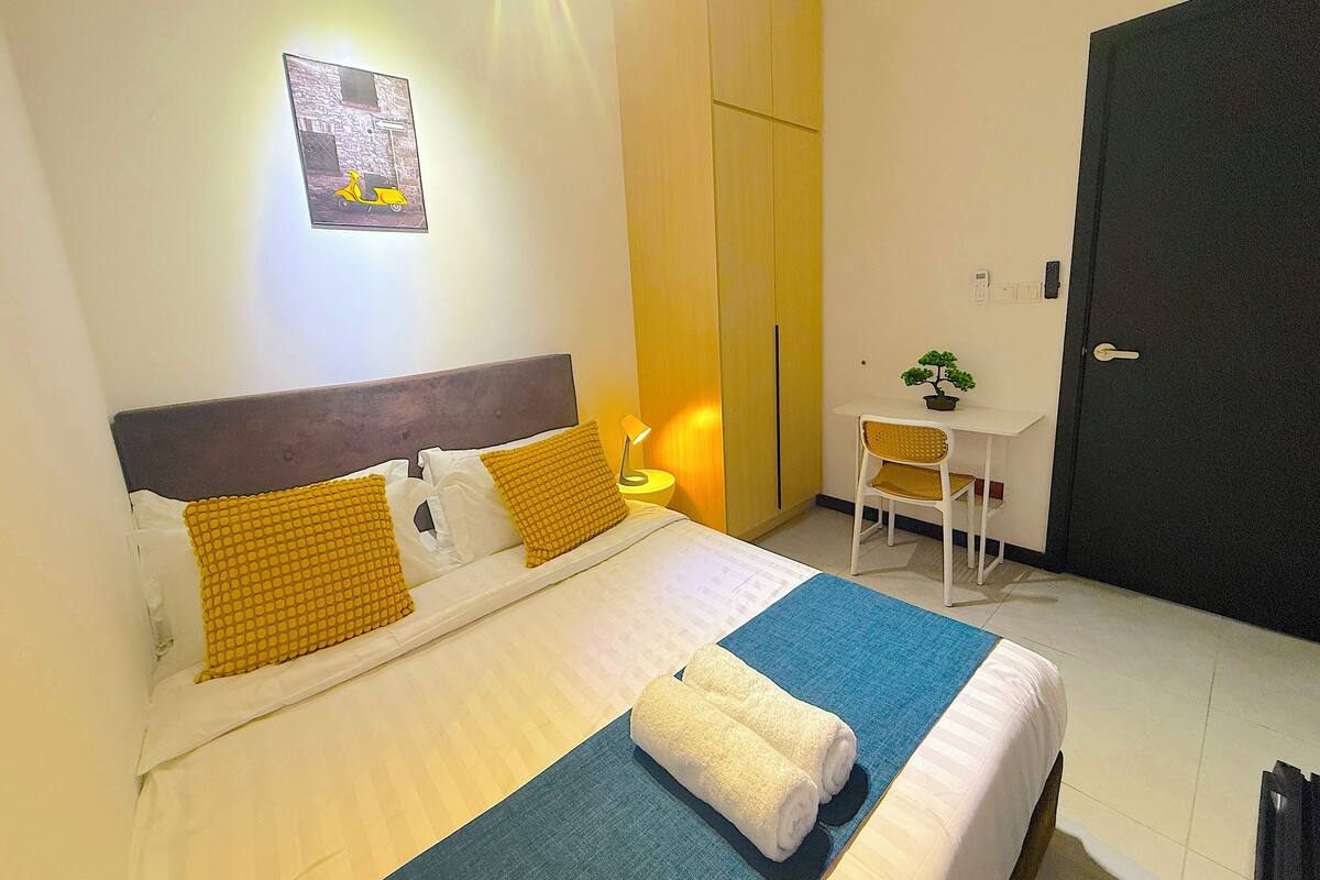 A neatly made bed with white and yellow bedding, a blue mat, and two rolled towels. There is a desk with a chair and a potted plant, a wardrobe, and a door.