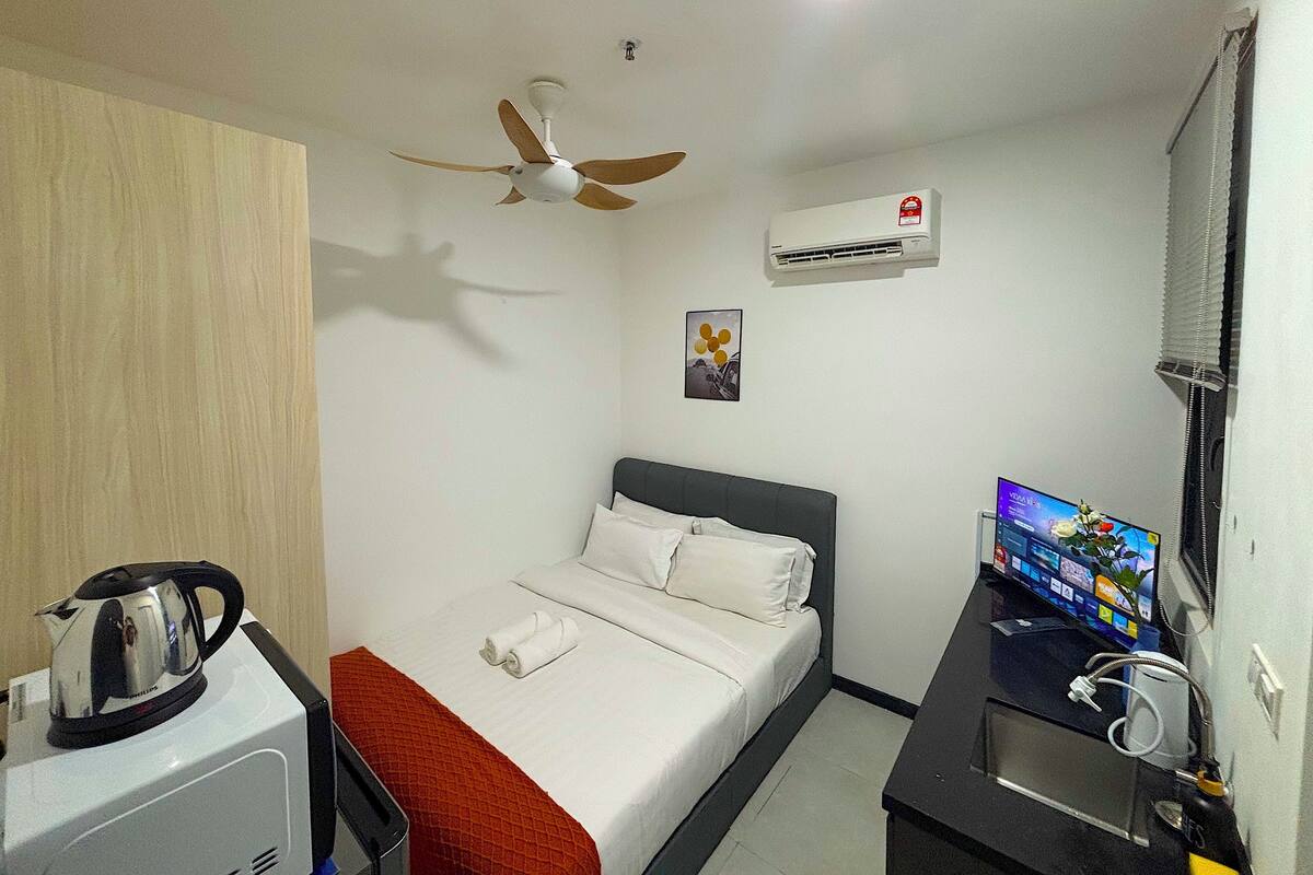 A hotel room with a bed, a ceiling fan, an air conditioning unit, a television, and a kettle on a small table.