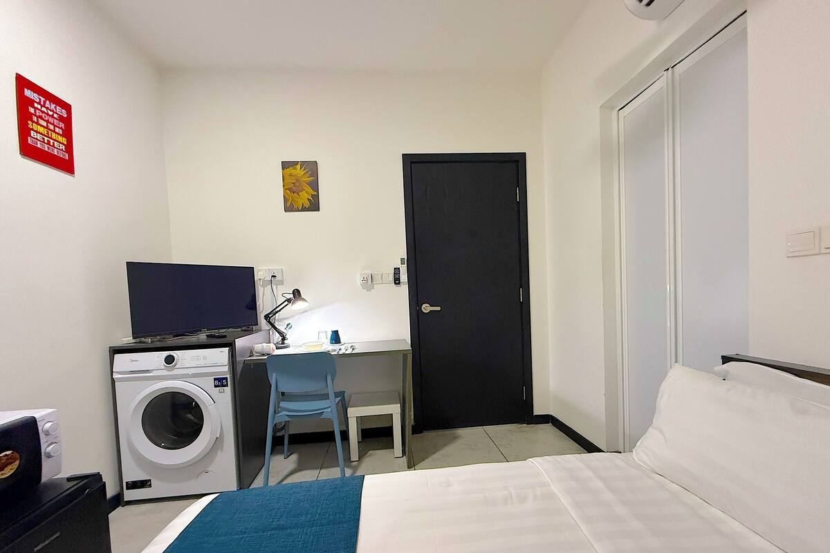 A hotel room with a bed, a desk with a chair, a washing machine, a television, and a door.